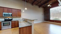 This 2-bedroom, 1-bathroom loft-style residence showcases a compelling blend of industrial heritage and contemporary design. The open-concept living and kitchen area is anchored by striking exposed wooden ceiling beams and visible ductwork, lending the space a distinctive character. Warm bamboo hardwood flooring flows throughout the main living areas, complementing the rich tones of the exposed brick accent walls.

The kitchen is well-appointed with rich cherry wood cabinetry, granite countertops, a breakfast bar peninsula with pendant lighting, and a full suite of stainless steel appliances including a side-by-side refrigerator, electric range with built-in microwave, and dishwasher. Soaring ceiling heights in the main living area create a dramatic sense of openness, with exposed industrial columns adding to the loft aesthetic.

The two bedrooms are carpeted for comfort, with one bedroom featuring a large picture window framed by exposed brick, offering views of the surrounding area. The full bathroom includes a tub-shower combination and granite-topped vanity with cherry cabinetry, and conveniently houses a stacked washer and dryer unit. Throughout the residence, clean white trim and neutral wall tones provide a polished contrast to the raw industrial elements.