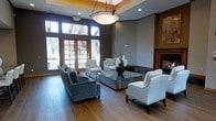 This refined single-floor space exudes a polished, welcoming ambiance through its thoughtful design and high-end finishes. Rich hardwood flooring anchors the room, complementing the warm wood trim that frames the large French doors and transom windows along one wall. These expansive windows flood the interior with natural light while offering views of the surrounding landscape, creating a bright and airy atmosphere.

The seating area is elegantly appointed with a plush blue-grey sofa and a pair of classic white accent chairs arranged around a sleek glass and metal coffee table, providing an inviting setting for conversation or relaxation. A built-in fireplace with a wood-trimmed surround serves as a distinguished focal point on one wall, adding warmth and sophistication to the space.

Overhead, a striking large pendant light fixture with a warm-toned tray ceiling detail draws the eye upward, enhancing the sense of vertical space and architectural character. Neutral grey walls provide a subtle backdrop that allows the furnishings and architectural elements to stand out. A decorative floral arrangement on the coffee table adds a finishing touch of elegance. Overall, the space strikes a balance between contemporary style and classic comfort, making it ideal as a communal lounge or gathering area.