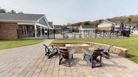 This inviting outdoor community space is thoughtfully designed to encourage relaxation and social gatherings. The grounds feature expansive natural stone paver hardscaping that seamlessly connects multiple distinct activity areas, creating a cohesive and polished outdoor environment.

At the heart of the space is a stone fire pit lounge area, encircled by a low stacked-stone retaining wall and surrounded by Adirondack-style chairs, offering a cozy and welcoming setting for residents to gather. Adjacent to the fire pit, a covered pavilion supported by classic brick columns and white structural beams provides a shaded retreat complete with picnic-style seating — ideal for outdoor dining or casual meetings. The pavilion's open-air design ensures excellent sightlines across the manicured green lawn.

Beyond the patio, a fenced resort-style pool area is visible, equipped with lounge chairs and shaded umbrellas, adding to the recreational appeal of the community. The surrounding landscape features well-maintained grassy open areas and a playground visible in the background, making this space suitable for residents of all ages.

The residential buildings in the backdrop showcase a mix of brick and board-and-batten siding with pitched rooflines, contributing to a cohesive neighborhood aesthetic. Rolling wooded hills frame the horizon, enhancing the natural, community-oriented ambiance of this versatile outdoor amenity space.