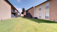 This single-story residence is part of a multi-building complex characterized by a mix of brick and vinyl siding exteriors. The grounds feature well-maintained, expansive lawns stretching between buildings, with concrete walkways, exterior staircases serving upper-level units, and HVAC equipment tucked neatly along the building's side. The outdoor environment is bright and open, benefiting from mature trees and generous green space shared across the property.

Inside, the interior presents a refined aesthetic with neutral, cream-toned walls and dark, polished concrete-style flooring that grounds the space with an industrial-modern sensibility. Recessed lighting illuminates the dining area, which serves as a focal point of the interior. A striking round marble-topped pedestal table in deep green anchors the room, surrounded by woven rattan chairs atop a natural fiber rug, lending warmth and texture to the space.

A large framed abstract artwork in soft, neutral tones adorns the wall, adding an artistic dimension. A sliding barn door on a black metal track offers a practical yet stylish room divider, connecting to an adjacent space where rustic antique-framed doors and gallery-style wall art are visible, enhancing the eclectic, curated character of the overall interior.