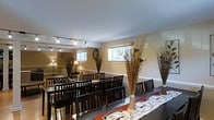 Located at 800 Kimberton Road in Phoenixville, Pennsylvania, this single-floor space is thoughtfully designed to accommodate gatherings and dining events. The main room features a series of long, dark-wood dining tables paired with matching dark slatted chairs, arranged to maximize seating capacity while maintaining an organized and refined aesthetic. Decorative arrangements of tall dried branches in floor vases add a natural, artistic touch to the space, complementing the warm, neutral tones of the walls. A wainscoting detail along the lower portion of the walls adds a classic architectural element, enhancing the overall elegance of the interior. Overhead, a track lighting system provides adjustable, even illumination throughout the room, while floor lamps in the corners contribute a softer, ambient glow. Multiple windows dressed with roman shades allow natural light to filter in, creating a bright and inviting atmosphere. A decorative wall clock serves as a subtle focal point, adding character to the room. The space is finished with area rugs that define the dining zones and add warmth underfoot. Overall, this venue offers a polished and versatile environment well-suited for private dining, community events, or organized gatherings.