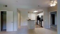 This single-floor residence offers a well-designed open-concept layout that seamlessly connects the living and kitchen areas. The kitchen is a functional centerpiece, equipped with stainless steel appliances including a refrigerator, gas range, and built-in microwave. White cabinetry provides ample storage, while a peninsula with a dark countertop and integrated sink serves as a natural divider between the kitchen and the adjoining living space. The kitchen features hardwood-style flooring, and a recessed skylight panel enhances brightness overhead. The living area is finished with plush carpeting and painted in neutral, soft tones that create a calm and welcoming atmosphere. A ceiling fan with a light fixture provides both comfort and illumination, while a window fitted with blinds allows for natural light and privacy control. A sliding panel door adds a clean, contemporary storage solution to the living space. A hallway leads to additional rooms beyond, suggesting a practical and well-organized floor plan. Throughout the space, clean lines, neutral color palettes, and consistent finishes contribute to a cohesive and move-in-ready environment.