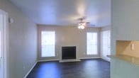This single-floor residence offers a comfortable and well-appointed living space designed with both style and functionality in mind. The living room serves as the heart of the home, showcasing rich, dark hardwood flooring that contrasts elegantly with the neutral, light-toned walls. A classic fireplace with a clean tile surround anchors the room, creating a natural focal point and a cozy atmosphere. Flanking the fireplace are two generously sized windows dressed with horizontal blinds, flooding the space with abundant natural light and offering views to the exterior. A ceiling fan with an integrated light fixture is centrally mounted overhead, providing both air circulation and supplemental lighting. The ceiling features a textured finish that adds subtle visual interest to the room. A paneled door on one side of the living area provides convenient access, contributing to the practical flow of the floor plan. The overall ambiance is clean, open, and move-in ready, with a neutral palette that offers a versatile backdrop for a variety of interior design styles.