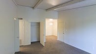 This single-floor residential space offers a clean, functional layout with a neutral palette throughout. The main living area is highlighted by distinctive exposed ceiling beams that add architectural character and a sense of height to the room. Soft, wall-to-wall carpeting in a warm gray tone runs throughout, providing comfort underfoot and a cohesive look across the space.

A built-in closet with a full-length hanging rod is seamlessly integrated into the wall, offering practical and organized storage. The open doorway leads into an adjoining room, also carpeted, which is bathed in warm light from a ceiling fixture, suggesting it functions as an additional bedroom or multipurpose room. A second door provides further access to other areas of the home, enhancing the flow and connectivity of the layout.

The walls are finished in a crisp, neutral white, complementing the light tones of the trim and doors throughout. Ventilation registers are visible, indicating a centralized climate control system. The overall ambiance is understated and versatile, offering a blank canvas that can be easily personalized to suit a variety of living arrangements. The space is well-suited for comfortable, everyday living with its straightforward, accessible design.