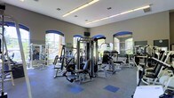 This fitness center offers a spacious and well-appointed workout environment designed to accommodate a comprehensive exercise routine. The facility is outfitted with an extensive selection of strength training machines, including a multi-station cable and weight machine system, as well as benches and free weight racks, providing options for both beginner and advanced users. The flooring features durable, rubberized material with blue accent tiles, offering both safety and a polished aesthetic. Large, arched windows line the walls, flooding the interior with abundant natural light and providing pleasant views of the surrounding outdoor greenery. Full-length mirrors are strategically placed along the walls, enhancing the sense of space and allowing users to monitor their form during workouts. The ceilings are notably high, contributing to an open and airy atmosphere throughout. Recessed linear lighting complements the natural light to ensure the space remains bright at all hours. The overall ambiance is clean, modern, and motivating, making this fitness center a functional and inviting space for daily physical activity.