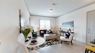 This single-floor residence offers a cohesive, contemporary design anchored by light wood-style flooring, crisp white walls, and elegant crown molding throughout. The open-concept living and dining area forms the heart of the home, where large windows invite generous natural light and provide outdoor views. The living space comfortably accommodates a full seating arrangement and is anchored by a bold patterned area rug, lending warmth and visual interest to the room.

The galley-style kitchen is thoughtfully designed with white shaker cabinetry, dark granite countertops featuring dramatic veining, a marble-style tile backsplash, and stainless steel appliances. A peninsula with bar seating and pendant lighting connects the kitchen to the entry and living areas, enhancing the open flow.

The bedroom is generously proportioned, featuring a walk-in closet, a window with outdoor views, and ample space for a full furniture suite. Crown molding continues into the room, maintaining a refined aesthetic throughout.

The bathroom is finished with dark granite vanity counters, white cabinetry, and a soaking tub set against marble-style tile surround, offering a clean, spa-inspired atmosphere. Overall, the residence presents a polished and functional living environment.