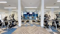 This fitness center offers a thoughtfully designed workout environment that combines functionality with contemporary aesthetics. The space features a bright, open layout supported by white structural columns that define distinct workout zones without compromising the overall sense of spaciousness. Natural and artificial lighting work in harmony, with linear pendant fixtures illuminating the floor throughout.

The flooring makes a strong visual statement, blending herringbone wood-look planks in the entry and transition areas with bold geometric patterned carpet tiles in blue, white, and gray tones across the main workout floor. A vibrant blue accent wall anchors the far end of the space, adorned with graphic circular design elements and serving as a dynamic focal point.

Cardio equipment, including multiple treadmills and elliptical machines, is arranged in organized rows, providing ample space between units for comfortable use. Multiple wall-mounted flat-screen televisions are positioned throughout, ensuring visibility from virtually any point in the facility. Additional equipment and strength training stations are visible toward the rear of the space. Large mirrors line the walls, enhancing the sense of depth and allowing users to monitor their form. The overall ambiance is energetic yet clean and modern, creating an inviting atmosphere for a full range of fitness activities.