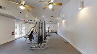 Located at 5500 SW Archer Rd in Gainesville, Florida, this fitness center offers a dedicated exercise space designed to support a well-rounded workout routine. The room features vaulted ceilings that lend an open, airy feel, complemented by multiple ceiling fans that ensure comfortable air circulation throughout. The flooring is covered in durable, grey carpet suited for fitness use. Natural light flows in through several windows and glass doors, creating a bright and welcoming environment. The space is equipped with a multi-station cable weight machine for strength training, along with cardio equipment including an elliptical trainer, a stationary upright bike, and a stair climber. Additional free weights and a weight bench are also present, offering versatility for various workout styles. Glass double doors provide easy access to the adjoining facility, and the clean white walls keep the space feeling fresh and open. Recessed and wall-mounted lighting supplements natural light for evening use. The layout allows for ample room to move between equipment, making it functional for multiple users simultaneously. This exercise room serves as a convenient and well-maintained amenity for residents of the surrounding community.