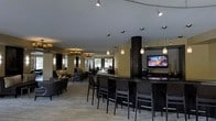 This single-floor space functions as a sophisticated lounge and bar area, thoughtfully designed to accommodate both casual gatherings and relaxed socializing. At its centerpiece is a curved bar counter finished in dark materials, lined with tall bar-height chairs that invite guests to gather around. A flat-screen television mounted nearby adds an entertainment focal point to the bar area, making it well-suited for watching events or casual viewing.

A striking dark mosaic column anchors the space visually, contributing to the upscale aesthetic. The ceiling features an eclectic mix of lighting solutions, including track lighting, elegant pendant lights in teardrop and cylindrical forms, and a traditional chandelier visible toward the rear — together creating layers of warm, ambient illumination throughout.

Beyond the bar, a comfortable lounge area is furnished with low-profile armchairs and sofas in dark upholstery, arranged to encourage conversation. The neutral-toned carpet and warm wall colors lend a welcoming, refined atmosphere. Large windows at the far end allow natural light to filter in, adding brightness and a sense of openness to the overall space.
