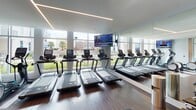 Located at 4307 Sharon Rd in Charlotte, NC, this modern fitness center delivers a comprehensive, well-appointed workout facility designed with both function and aesthetics in mind. The space is characterized by rich hardwood-style flooring, crisp white walls, and striking linear LED lighting that runs across the ceilings in geometric patterns, creating a clean, energetic atmosphere throughout.

The cardio zone is a standout feature, lined with a row of state-of-the-art treadmills and stationary bikes, including Peloton cycling stations, all positioned to take full advantage of the expansive floor-to-ceiling windows. These large windows flood the space with abundant natural light and offer open views of the surrounding outdoor greenery and neighborhood beyond.

The strength training area is generously equipped with a wide array of commercial-grade cable machines, resistance equipment, dedicated benches, and a medicine ball rack, providing options for a full-body workout. Large mirrors along the walls enhance the sense of space and support proper form during training. Overhead televisions are mounted for entertainment during cardio sessions.

The entry area features glass-panel doors, built-in shelving, and a clean, organized welcome space. The overall ambiance is polished and professional, reflecting a high-end fitness environment suited for a variety of training needs.