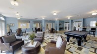 This single-floor clubhouse is thoughtfully designed to serve as a premier gathering and recreational destination. The open-concept layout is anchored by a comfortable lounge area furnished with plush leather seating, including a sofa and accent chairs arranged around a round ottoman coffee table, creating an inviting space for relaxed conversation. A bold geometric-patterned area rug in neutral tones of green, beige, and gray anchors the seating arrangement and adds a striking visual element to the interior.

Adjacent to the lounge, a full-size billiards table provides a focal point for recreation and entertainment. The space also features multiple dining areas with bistro-style tables and chairs, ideal for casual gatherings or events. Soft blue-gray walls complement the light-toned flooring throughout, giving the clubhouse a calm and contemporary ambiance.

French-style glass doors line the perimeter, allowing natural light to filter in and offering views of the surrounding exterior. Flush-mount ceiling fixtures provide consistent, warm illumination throughout the space. A cue rack and additional game furnishings are visible along the walls, further reinforcing the recreational character of the clubhouse. Overall, the space balances comfort, function, and style in a well-appointed community setting.