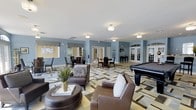This single-floor clubhouse is thoughtfully designed to serve as a premier gathering and recreational destination. The open-concept layout is anchored by a comfortable lounge area furnished with plush leather seating, including a sofa and accent chairs arranged around a round ottoman coffee table, creating an inviting space for relaxed conversation. A bold geometric-patterned area rug in neutral tones of green, beige, and gray anchors the seating arrangement and adds a striking visual element to the interior.

Adjacent to the lounge, a full-size billiards table provides a focal point for recreation and entertainment. The space also features multiple dining areas with bistro-style tables and chairs, ideal for casual gatherings or events. Soft blue-gray walls complement the light-toned flooring throughout, giving the clubhouse a calm and contemporary ambiance.

French-style glass doors line the perimeter, allowing natural light to filter in and offering views of the surrounding exterior. Flush-mount ceiling fixtures provide consistent, warm illumination throughout the space. A cue rack and additional game furnishings are visible along the walls, further reinforcing the recreational character of the clubhouse. Overall, the space balances comfort, function, and style in a well-appointed community setting.