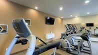 This fitness center offers a focused and functional workout environment thoughtfully designed for cardio training and general exercise. The space features a selection of cardio equipment, including elliptical machines and a recumbent bike, arranged to allow comfortable use and easy movement between machines. A wall-mounted flat-screen television is positioned for visibility from the equipment, providing entertainment during workouts. The mirrored wall along one side of the room enhances the sense of space while allowing users to monitor their form. Recessed lighting in the smooth ceiling provides bright, even illumination throughout the room, contributing to an energized atmosphere. The warm-toned walls create a welcoming environment balanced by the clean, modern aesthetic of the equipment and flooring. A wall-mounted climate control unit ensures a comfortable temperature during workouts. Small utility shelves are also present on the wall, offering convenient storage for personal items. The single-floor layout ensures easy accessibility, making this a practical and inviting fitness facility suited for daily use.