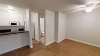 This single-floor space offers a straightforward, functional layout designed for comfortable everyday living. The open floor plan connects the kitchen and living area seamlessly, creating a cohesive and accessible flow throughout the unit. The kitchen is positioned to one side and features dark countertops that contrast neatly against white cabinetry and appliances, including a full-size refrigerator. Hardwood-style flooring runs throughout the main living areas, providing a warm and durable surface underfoot. The walls are finished in a neutral light gray tone, contributing to a clean, modern aesthetic that allows for versatile interior styling. A textured ceiling adds a subtle architectural detail across the space. A bathroom is accessible directly from the main living area, featuring standard fixtures in a compact, well-organized layout. Doorways and open passageways between spaces create a sense of depth and connectivity throughout the floor plan. The overall ambiance is bright, neutral, and move-in ready, making it a practical and welcoming space suitable for a range of occupants.