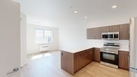 This thoughtfully designed 1-bedroom, 2-bathroom residence spans a single floor and makes excellent use of its well-proportioned layout. The open-concept kitchen and living area serves as the heart of the home, featuring warm wood-toned cabinetry, a large peninsula with a white quartz countertop, and a full suite of stainless steel appliances including a refrigerator, range, microwave, and dishwasher. Recessed lighting and light hardwood flooring run throughout, lending the space a clean, contemporary aesthetic. The living area receives generous natural light through a wide window with neighborhood views, complemented by a wall-mounted heating unit for year-round comfort.

The bedroom is a bright, comfortable retreat with its own window overlooking the surrounding neighborhood, a ceiling fan, and light hardwood floors that continue from the main living space. A dedicated walk-in closet with built-in wire shelving provides practical storage. Two bathrooms serve the unit, one of which features a full soaking tub and shower combination with clean white tiled surround and accessibility grab bars. A hallway connects the private areas of the residence, completing this efficient and modern floor plan.