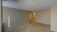 This single-floor residence offers a straightforward and functional layout designed to maximize usable living space. The interior is finished with wall-to-wall carpeting in a warm neutral tone, providing a soft and comfortable underfoot experience throughout the main living areas. The walls are painted in a consistent light beige, creating a clean and cohesive aesthetic that serves as a versatile backdrop for a variety of décor styles. The textured popcorn ceiling adds a classic residential character to the space, while a ceiling fan in the adjoining area provides both practical air circulation and a subtle lighting element. Visible HVAC vents are integrated into both the ceiling and walls, ensuring climate control throughout the space. The open layout allows for a natural flow between the main living zone and the adjacent room, which is defined by a partial dividing wall and railing detail, adding a gentle architectural transition between spaces. The neutral palette, combined with the open connectivity between rooms, creates an inviting atmosphere that is well-suited for comfortable everyday living. Overall, the space presents a clean, move-in-ready environment with classic finishes and a functional design.