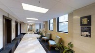 This single-floor space presents a polished, professional corridor that doubles as a reception or waiting area. The hallway is finished with light-toned wall cladding — likely stone or composite panels — lending a refined, institutional character to the interior. The flooring features a classic checkered tile pattern in neutral tones, contributing to a clean and orderly appearance.

Overhead, a suspended drop ceiling with large recessed fluorescent light panels ensures consistent, bright illumination throughout the corridor. A row of double-hung windows along one wall allows natural light to filter in, providing views of an adjacent exterior structure and enhancing the overall brightness of the space.

The seating area features mid-century modern style armchairs with wood frames and soft upholstered cushions in a muted blue-grey tone, arranged along the window side for a comfortable waiting experience. Potted indoor plants are thoughtfully placed throughout, softening the architectural lines and adding a touch of greenery. At the far end of the corridor, wooden doors provide access to additional areas, rounding out a functional and well-maintained space.