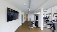 This single-floor fitness facility presents a clean, modern aesthetic characterized by crisp white walls and smooth, durable flooring in a warm neutral tone. The space is thoughtfully designed to accommodate a range of exercise activities, with a layout that maximizes usability and flow. A large wall-mounted flat-screen television is positioned prominently on one wall, providing entertainment or instructional content during workouts. The ceiling features recessed lighting alongside dedicated overhead fixtures, ensuring the space is well-lit throughout. A multi-functional cable resistance machine serves as a centerpiece of the strength training area, complemented by additional equipment visible toward the far end of the room, suggesting a broader selection of cardio and strength options. Structural columns punctuate the space while defining distinct workout zones. A secured exit door is visible toward the rear, contributing to the room's safety infrastructure. Built-in speakers in the ceiling further enhance the workout environment. The overall atmosphere is clean, professional, and purpose-built, reflecting a well-maintained and fully equipped fitness space.