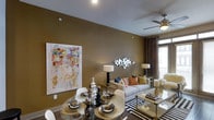 This single-floor space showcases a thoughtfully designed open-concept layout that combines the dining and living areas into one cohesive, flowing environment. The walls are finished in a rich, earthy olive-gold tone that lends warmth and depth to the interior, complemented by light gray ceiling panels that add a subtle architectural contrast.

The dining area features a round glass-top table surrounded by modern white chairs, set atop woven placemats and styled with elegant tableware, creating a refined yet approachable atmosphere. A large, vibrant abstract canvas dominates the adjacent wall, introducing bold splashes of color that animate the space.

The living area is anchored by a plush gray sofa dressed in an array of colorful accent pillows in orange, blue, and gold. A geometric mirrored wall sculpture adds a decorative focal point, while a ceiling fan with integrated lighting provides both function and style. Gold-toned glass side tables and a striped area rug complete the contemporary aesthetic.

Natural light filters in through draped windows, enhancing the warm ambiance throughout. The overall design strikes a balance between sophistication and comfort, with carefully curated furnishings and décor that give the space a polished, magazine-ready character.