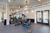 This single-level fitness center is thoughtfully designed to support a comprehensive workout experience across multiple distinct zones. The space is finished with durable rubber flooring throughout the main exercise areas, transitioning to light hardwood-style flooring in the open studio section, and is illuminated by a combination of recessed lighting and stylish pendant fixtures paired with ceiling fans for comfort.

The strength training area is equipped with a full selection of selectorized machines, a dual cable cross station, a dumbbell rack with adjustable benches, and large wall-mounted mirrors that enhance the sense of space. A motivational quote — "If it doesn't challenge you, it won't change you" — is prominently displayed along the upper wall, reinforcing the facility's active atmosphere. Brick-accented columns add an architectural detail that grounds the modern design aesthetic.

The cardio zone features treadmills, ellipticals, and a cycling bike, positioned near floor-to-ceiling windows that invite abundant natural light and outdoor views. A separate, open-plan studio area provides ample space for group exercise, yoga, or functional training, and is stocked with resistance equipment, stability balls, and a smart training system. Glass-paned double doors connect the main gym floor to additional areas, ensuring seamless flow throughout the facility.