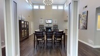 Located at 1 Westchase Dr in Charleston, SC 29407, this single-floor community space is defined by its dramatic double-height ceilings and an expansive open-concept layout that seamlessly connects the living, dining, and kitchen areas. A continuous row of clerestory windows encircles the upper walls of the main gathering area, flooding the interior with natural light and creating an airy, uplifting atmosphere throughout the day.

The main living area is anchored by a classic fireplace flanked by open shelving, and furnished with generous cream-toned sofas arranged around a dark wood coffee table. A wall-mounted flat-screen television and carefully selected artwork add to the refined yet welcoming character of the space. An elegant chandelier with a contemporary design hangs overhead, complementing the rich, dark hardwood flooring that runs throughout.

Adjacent to the living area, the dining space accommodates a large dark wood high-top table with seating for several guests, making it ideal for gatherings and events. Decorative cabinetry and accent pieces complete the room's polished aesthetic.

The kitchenette is outfitted with marble-patterned granite countertops, a stainless steel refrigerator, an undermount sink, and recessed lighting, providing convenient service amenities. French doors offer views of and access to the surrounding greenery, further enhancing the connection between the interior and the natural landscape outside.