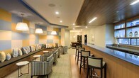 This single-floor communal lounge space is thoughtfully designed to serve as a vibrant social hub. The interior immediately draws attention with its eye-catching geometric wall accents in alternating blue and orange panels, adding a modern, playful character to the environment. Recessed lighting throughout the ceiling is complemented by elegant pendant lights, creating a warm and welcoming ambiance.

The layout is divided into distinct functional zones. A built-in cushioned banquette runs along one wall, flanked by patterned armchairs and small side tables, forming a comfortable lounge area ideal for casual conversation. The neutral-toned carpeting in this seating zone adds softness underfoot, while hardwood flooring in the adjacent areas enhances the transition between spaces.

A prominent bar counter with high-top bar stools stretches along one side of the room, providing a dedicated area for socializing or enjoying refreshments. The bar area features a light blue countertop that ties in with the room's color palette. Toward the rear of the space, a billiards table serves as a central entertainment feature, rounding out the recreational offerings. A wood-paneled ceiling detail in the bar and game area adds a rich, contrasting texture that elevates the overall design aesthetic.