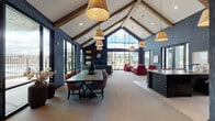 Located in Beaverton, Oregon, this single-story community clubhouse on SW 172nd Terrace delivers a striking blend of contemporary design and inviting communal spaces. The architecture is defined by a dramatic vaulted ceiling with exposed wooden beams that rise to a peak, anchored by a wall of floor-to-ceiling windows that floods the main gathering area with natural light and provides views of the surrounding community and outdoor amenity spaces.

Throughout the interior, deep navy textured wall coverings establish a bold, sophisticated atmosphere, contrasted by neutral carpeted flooring and crisp white ceiling panels. The main open-plan space accommodates a large dining table with upholstered seating and an adjacent lounge area furnished with bold crimson armchairs and a low-profile coffee table, creating distinct zones for dining and casual conversation.

A well-appointed kitchen features dark cabinetry, a range with a wall-mounted hood, open display shelving, and a central island with light wood countertops and bar-height seating. A separate lounge nook offers a built-in fireplace flanked by dark shelving, large windows overlooking the outdoor pool area, and intimate seating. Pendant lighting throughout ties the design together, reinforcing the space's polished, modern character.