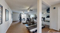 Located at 3201 SW Twilight Ct in Topeka, Kansas, this single-floor fitness center offers a comprehensive workout facility designed to support a variety of exercise routines. The gym is outfitted with a well-rounded selection of cardio equipment, including treadmills, elliptical trainers, and a recumbent stationary bike, all positioned on protective floor mats over wall-to-wall carpeting. A multi-station cable weight machine anchors the strength training area, complemented by a weight bench and free weights for a full range of resistance exercises. Large wall-mounted mirrors span a significant portion of the space, visually expanding the room and allowing users to monitor their form during workouts. A wall-mounted television provides entertainment during exercise sessions. Multiple ceiling fans are distributed throughout the room, ensuring consistent airflow and comfort. Natural light filters in through window treatments along the exterior wall, lending a bright and welcoming atmosphere to the space. The textured ceiling and neutral-toned walls contribute to a clean, functional aesthetic. An adjacent hallway with carpeted flooring connects to a small sitting area, adding a comfortable transitional space just outside the main gym area.