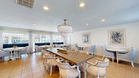 Located in Virginia Beach, VA, this well-designed community room offers a welcoming and versatile gathering space finished in a coastal-inspired palette of soft blues, whites, and warm neutrals. The main dining area is anchored by a substantial rustic wood farmhouse table surrounded by upholstered chairs, all illuminated by a striking beaded chandelier with gold accents that serves as the room's centerpiece. Recessed lighting throughout ensures a bright, inviting atmosphere.

Multiple intimate seating arrangements with round pedestal tables and cushioned armchairs are thoughtfully positioned around the room, offering flexible spaces for smaller gatherings. Large windows dressed with sheer white curtains and blinds flood the interior with natural light and provide views to the outdoors. Coastal-themed framed artwork adorns the walls, enhancing the relaxed, seaside ambiance.

The fully equipped kitchen features crisp white shaker-style cabinetry, white countertops, and stainless steel appliances including a range, microwave, dishwasher, and refrigerator, along with a convenient peninsula for additional prep or serving space. Warm tile flooring flows consistently throughout, tying the entire space together into a cohesive, comfortable environment ideal for community events and social gatherings.