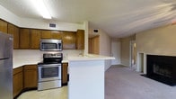 This single-floor residence offers a practical and comfortable layout designed for everyday living. The kitchen is a standout feature, outfitted with stainless steel appliances including an electric range and an over-the-range microwave, complemented by warm-toned wood cabinetry that provides ample storage. A white peninsula with a built-in sink doubles as a breakfast bar, seamlessly connecting the kitchen to the adjacent living area and creating an open, airy feel. The transition from tile flooring in the kitchen to plush carpeting in the living space adds a sense of distinction between functional and relaxing zones. The textured ceiling and recessed lighting contribute to the interior's understated, practical aesthetic. A hallway visible from the main living area leads to additional rooms, including what appears to be a bedroom and bathroom, suggesting a well-organized floor plan. The neutral color palette throughout — soft whites and warm beiges — creates a clean and inviting atmosphere. HVAC vents are integrated throughout the ceiling, ensuring year-round climate comfort. Overall, this space offers a cohesive and functional living environment suited for comfortable day-to-day life.
