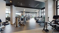 Located at 525 NE First Ave in Fort Lauderdale, Florida, this contemporary fitness center offers a well-appointed, multi-zone workout facility designed for a full range of exercise needs. The space features a sleek, modern aesthetic characterized by a bold dark ceiling contrast against crisp white walls, durable rubberized flooring, and floor-to-ceiling windows that flood the interior with natural light and frame views of the outdoor pool area and tropical palm tree surroundings.

The main workout floor is generously proportioned and organized into distinct functional zones. A dedicated strength training area houses an extensive selection of cable machines, multi-use weight stations, and a full rack of dumbbells alongside adjustable benches. A separate free weights section provides kettlebells and medicine balls neatly arranged on wall-mounted racks. The cardio zone features a long row of treadmills, elliptical trainers, and stair climbers positioned to take advantage of the outdoor views through the large windows.

A dedicated spin studio with hardwood flooring, wall-to-wall mirrors, and a mounted flat-screen display offers an immersive cycling experience. Additional amenities include locker facilities visible from the main floor. Throughout, large mirrors amplify the sense of space and provide functional workout support. Vibrant mural art adds a dynamic, energetic touch to the overall atmosphere.