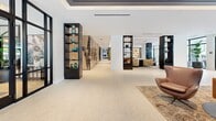 This generously sized clubroom in Sweetwater, Florida, delivers a sophisticated community gathering space defined by contemporary design and an open, airy layout. Light-toned stone tile flooring runs throughout, lending a clean and cohesive aesthetic that unifies the various zones within the space. Floor-to-ceiling windows with sleek black frames line the exterior walls, flooding the interior with abundant natural light and framing views of a palm-lined outdoor terrace, which is directly accessible through multiple glass doors. The terrace features additional seating, extending the usable social space outdoors. Inside, the clubroom is organized into distinct yet flowing lounge areas, each thoughtfully furnished with a curated mix of leather seating, curved sofas, and textured accent chairs arranged atop geometric patterned rugs. Dark, illuminated display columns punctuate the open floor plan, adding visual interest and serving as elegant dividers between zones. A central seating area anchors the space beneath a bold pendant light and a coffered ceiling with deep blue-toned panels, creating an intimate focal point. Large-scale artwork and a wall-mounted display screen add character, while lush indoor plants introduce natural warmth. The overall ambiance is polished, welcoming, and well-suited for both social gatherings and relaxed lounging.