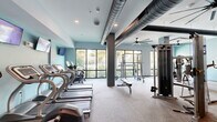 Located at 2201 NW Federal Highway in Stuart, Florida, this well-appointed fitness center offers a generously sized, single-floor layout designed to accommodate a full range of workout activities. The space is defined by its modern industrial aesthetic, with exposed metallic ductwork running across the ceiling, recessed lighting, and large ceiling fans that ensure continuous airflow throughout the facility.

Floor-to-ceiling windows line the perimeter, flooding the interior with natural light and providing open views of the surrounding tropical landscaping, including lush palm trees. The cardio zone features a row of treadmills and stationary bikes positioned directly in front of these windows, creating an energizing environment for aerobic workouts.

The strength training area is equally well-equipped, housing a squat rack with weight plates, a multi-function cable machine, adjustable benches, and a full dumbbell rack ranging across various weights. Wall-mounted televisions are positioned throughout, offering entertainment during workouts. Full-length mirrors in the strength zone allow for proper form monitoring.

The flooring transitions between carpet and hardwood, delineating different workout zones within the space. A dedicated entry area includes built-in bench seating and access to restroom facilities, completing the functional design of this comprehensive fitness facility.