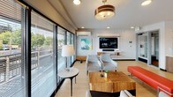 Located on North Proctor Street in Tacoma, Washington, this single-floor community lounge offers a thoughtfully designed, open-concept gathering space that seamlessly blends comfort with modern style. Light hardwood flooring flows throughout, unifying the distinct functional zones and contributing to a warm, inviting atmosphere.

The living area is generously proportioned, furnished with contemporary seating arrangements including upholstered chairs, a solid wood coffee table, and accent pieces. A wall-mounted flat-screen television with a built-in media shelf serves as the focal point, while large floor-to-ceiling sliding glass doors flood the space with natural light and provide direct views of the surrounding neighborhood streetscape. A wall-mounted mini-split system ensures year-round climate comfort.

The fully equipped kitchen is a standout feature, anchored by a central island with a built-in sink and seating for multiple guests. Stainless steel appliances — including a double wall oven, refrigerator, and range with a ventilation hood — line the perimeter. White upper cabinetry contrasts with warm wood lower cabinets, while open wooden shelving and a white subway tile backsplash add texture and visual interest. An exposed brick accent wall introduces an industrial touch, balancing the otherwise clean, contemporary aesthetic. Pendant lighting overhead enhances both function and ambiance throughout the kitchen zone.
