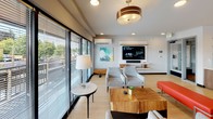 Located on North Proctor Street in Tacoma, Washington, this single-floor community lounge offers a thoughtfully designed, open-concept gathering space that seamlessly blends comfort with modern style. Light hardwood flooring flows throughout, unifying the distinct functional zones and contributing to a warm, inviting atmosphere.

The living area is generously proportioned, furnished with contemporary seating arrangements including upholstered chairs, a solid wood coffee table, and accent pieces. A wall-mounted flat-screen television with a built-in media shelf serves as the focal point, while large floor-to-ceiling sliding glass doors flood the space with natural light and provide direct views of the surrounding neighborhood streetscape. A wall-mounted mini-split system ensures year-round climate comfort.

The fully equipped kitchen is a standout feature, anchored by a central island with a built-in sink and seating for multiple guests. Stainless steel appliances — including a double wall oven, refrigerator, and range with a ventilation hood — line the perimeter. White upper cabinetry contrasts with warm wood lower cabinets, while open wooden shelving and a white subway tile backsplash add texture and visual interest. An exposed brick accent wall introduces an industrial touch, balancing the otherwise clean, contemporary aesthetic. Pendant lighting overhead enhances both function and ambiance throughout the kitchen zone.