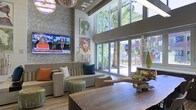 This dynamic single-story space makes a striking impression through its thoughtful blend of architectural scale and curated design. The room's most commanding feature is its double-height ceiling, which creates an airy, open atmosphere accentuated by exposed wood beam detailing and a sculptural chandelier that draws the eye upward.

A textured stone accent wall serves as a dramatic backdrop for a wall-mounted flat-screen television, grounding one end of the room with visual weight and warmth. The seating area is anchored by a contemporary sectional sofa in neutral tones, enlivened by boldly colored accent pillows in burnt orange and teal. Artisanal ceramic stools and a rustic driftwood-style coffee table introduce organic elements that complement the space's eclectic character.

Large-scale, colorful artwork adds personality throughout, contributing a gallery-like quality to the interior. Floor-to-ceiling sliding glass doors flood the space with abundant natural light while framing views of palm trees and a landscaped outdoor area beyond. A substantial wood table at the forefront suggests a versatile, social atmosphere suited for gathering and collaboration.