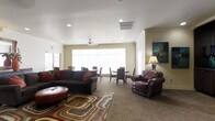 This single-floor space offers a well-appointed common area that seamlessly blends a lounge and dining zone into one cohesive, open layout. The lounge area is anchored by a large, dark sectional sofa paired with a rich leather armchair and a round leather ottoman, all arranged atop a colorful geometric area rug that adds warmth and visual interest to the carpeted floor. Recessed lighting and a ceiling fan keep the space bright and comfortable, while large windows along the perimeter flood the room with abundant natural light and contribute to an open, airy atmosphere. Soft, neutral wall tones in cream and warm beige provide a calm backdrop that complements the rich, earthy furnishings. A tasteful piece of artwork adds a touch of color to one wall, lending the space a curated, welcoming character. Toward the rear, a dining area features a set of tables and chairs, making the space functional for both casual gatherings and seated meals. The overall ambiance is relaxed yet purposeful, designed to accommodate a variety of social activities in a comfortable, communal setting.
