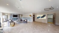 This well-appointed residential lobby presents a seamless blend of functionality and upscale design across a single, open floor. The herringbone-patterned light wood flooring extends throughout the entire space, creating a cohesive and refined aesthetic. At the heart of the lobby is a curated lounge area furnished with plush velvet armchairs, a rounded coffee table atop a textured area rug, and a wall-mounted flat-screen television set against a dramatic marbled accent wall featuring a built-in linear fireplace. Adjacent to the lounge, a full wall of individual mailboxes provides residents with convenient mail access, framed by subtly textured neutral wallcovering that adds warmth and depth to the space. The elevator vestibule features two stainless steel elevator doors set against coordinating textured walls, with recessed lighting above that keeps the area bright and welcoming. A separate seating nook benefits from an abundance of natural light, with floor-to-ceiling windows offering open views of the surrounding neighborhood streetscape and landscaping. Recessed lighting throughout the lobby ensures consistent, comfortable illumination. The overall ambiance is contemporary and polished, reflecting a thoughtfully designed communal environment that balances elegance with everyday practicality.