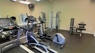 This fitness center is a dedicated single-floor exercise space designed to support a well-rounded workout routine. The room is outfitted with a selection of cardio equipment, including treadmills, elliptical trainers, and a stationary cycle, providing ample options for aerobic exercise. A cable resistance machine is positioned along one wall, offering strength and resistance training capabilities. The flooring consists of dark rubber matting, a practical and durable surface well-suited for a high-use fitness environment. Full-length mirrors line the walls, visually expanding the space while allowing users to monitor their form during exercise. Overhead fluorescent lighting ensures the room is well-lit throughout. A ceiling fan is mounted above, contributing to air circulation during workouts. The walls are finished in a neutral tone, keeping the atmosphere clean and functional. The overall layout is efficiently organized, making good use of the available floor space to accommodate multiple users and equipment types simultaneously. This fitness center provides a practical and accessible setting for residents or users seeking convenient on-site exercise facilities.