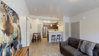 This single-floor residential unit offers a thoughtfully designed open-concept layout that seamlessly connects the living, dining, and kitchen areas. The living room features light wood-look flooring, a tufted sofa, and a bright window that invites natural light. A patterned area rug and clean white walls give the space a fresh, welcoming feel.

The kitchen is a standout feature, equipped with stainless steel appliances — including a refrigerator, microwave, and range — complemented by rich, dark wood cabinetry with modern hardware and light countertops. A peninsula with bar seating and pendant lighting above serves as both a functional workspace and casual dining spot, while a separate high-top dining table provides additional seating nearby. Track lighting enhances the kitchen's bright, open atmosphere.

The unit includes two well-sized bedrooms, both carpeted for comfort. The primary bedroom is particularly spacious, featuring a walk-in closet with built-in shelving and natural light from a high window. An en-suite bathroom offers a shower-tub combination with light wood-look flooring. The second bedroom also benefits from natural light through full-length blinds and a transom window. An in-unit washer is tucked conveniently within the bathroom area, adding everyday practicality to this well-rounded living space.