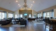 This expansive single-floor space is defined by its striking architectural design, most notably the soaring vaulted ceilings that lend an open, airy quality to the interior. A grand chandelier serves as a central focal point overhead, complemented by recessed lighting that ensures the space is well-illuminated throughout.

The room's most commanding feature is a floor-to-ceiling stacked stone accent wall, which anchors the space with a natural, textural contrast against the neutral-toned walls. A flat-screen television is mounted within this feature wall, adding a functional element to the dramatic backdrop.

Rich, dark hardwood flooring extends throughout, grounding the space and providing warmth. Multiple seating arrangements, composed of contemporary dark upholstered sofas and armchairs paired with round wooden coffee tables, are organized across area rugs, creating distinct yet open conversation zones.

Large windows along the perimeter allow natural light to filter in from multiple directions, enhancing the sense of openness. The overall ambiance balances elegance with comfort, making it well-suited as a communal gathering or lounge area.