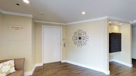 This single-floor lobby offers a clean, well-appointed entryway designed to make a strong first impression. The space is finished with warm cream-toned walls that lend a sense of brightness and openness, complemented by crisp white crown molding that frames the room with a classic architectural touch. The flooring features wood-look planks laid in a herringbone-style pattern, adding warmth and visual interest underfoot. A white paneled entry door serves as the focal point of the space, framed neatly within a clean white surround. Recessed lighting in the textured white ceiling provides even, ambient illumination throughout. A standout decorative element is the ornate scrollwork wall medallion displayed prominently on one wall, adding an artistic flourish that elevates the overall aesthetic. Wide white baseboards complete the refined look, tying together the neutral color palette. The overall ambiance is polished yet welcoming, balancing functional simplicity with thoughtful decorative details that set the tone for the space beyond.