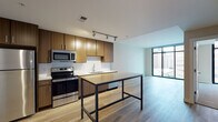 Located in the heart of Cincinnati, Ohio, this contemporary single-floor residence at 151 Fifth St W offers a sleek, modern aesthetic with thoughtful finishes throughout. The open-concept kitchen is a standout feature, equipped with stainless steel appliances including a refrigerator, range, microwave, and dishwasher. Warm wood-toned cabinetry with clean-lined hardware pairs elegantly with white quartz countertops and a classic subway tile backsplash. Light hardwood flooring and track lighting complete the kitchen's polished look, while a central island provides additional prep and dining space. The living and bedroom areas are carpeted for comfort and benefit from dramatic floor-to-ceiling windows framed in sleek black, flooding the space with natural light and offering sweeping views of the downtown Cincinnati skyline. The bathroom is well-appointed with a wood-tone vanity, white countertop, an undermount sink, a large mirror, and a tiled tub-shower combination. The overall ambiance of the space is clean, contemporary, and bright, with an urban edge that makes the most of its prime downtown location.