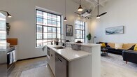 This single-floor unit showcases a thoughtfully designed industrial-modern aesthetic, defined by soaring ceilings, exposed mechanical elements, and large steel-framed windows that flood the interior with abundant natural light. The open-plan layout seamlessly connects the kitchen, dining, and living areas, creating a cohesive and airy atmosphere throughout.

The kitchen is a standout feature, centered around a generous island with a white quartz waterfall countertop, an undermount sink, and a tiled accent panel. Stainless steel appliances—including a range with a hood, refrigerator, and dishwasher—are complemented by white cabinetry and open shelving with curated greenery. Pendant lights and track lighting add warmth overhead.

The living area offers an open, comfortable layout with tall ceilings and polished concrete floors that carry throughout the unit, reinforcing the industrial character. The bedroom is a calm retreat featuring tall walls, a black-framed window with a roller shade, and a built-in closet with dark sliding doors. The bathroom maintains the modern edge with polished concrete floors, a glass-enclosed walk-in shower with white subway tile, and clean white fixtures accessed through a matte dark door.