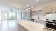 This thoughtfully designed single-floor residence showcases a contemporary aesthetic defined by clean lines, a neutral palette, and high-quality finishes throughout. The open-concept kitchen serves as the centerpiece, featuring a generous waterfall-edge quartz island with subtle veining, complemented by a pendant light fixture with brass accents. Cabinetry combines matte gray lower cabinets with warm wood-toned uppers and open shelving, anchored by a textured tile backsplash. A gold-finish faucet, undermount sink, and full suite of stainless steel appliances—including a range, dishwasher, refrigerator, and microwave—complete the space. The kitchen flows seamlessly into the living area, where light-toned herringbone hardwood flooring adds visual interest. The primary bedroom is generously proportioned, featuring a large window with city and treetop views and a ceiling fan. It connects to a walk-in closet with adjustable shelving systems and a well-appointed en-suite bathroom with a dual-sink vanity in warm wood tones, white quartz countertops, large-format tile flooring, and a glass-enclosed shower. A secondary bathroom mirrors the same refined finishes. An in-unit washer and dryer add everyday convenience to this modern residence.