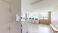 Located at 2775 Jane St in Toronto, Ontario, this shared laundry facility offers a practical and well-organized space designed to serve building residents efficiently. The facility is composed of two laundry rooms and an entrance area, all situated on a single level.

The laundry rooms are generously sized and feature durable speckled terrazzo flooring that is easy to maintain and clean. Warm beige walls create a neutral and functional atmosphere throughout, while fluorescent overhead lighting ensures the space is well-lit. Both rooms are equipped with multiple commercial-grade front-loading washing machines and top-loading dryers, arranged in neat rows along the walls to maximize usable floor space. Faucets and utility connections are mounted directly above the washers for convenient access.

A centrally placed folding station with a dark countertop provides a dedicated surface for sorting and folding laundry. A wooden bench along one wall offers a comfortable place to wait. Informational notices and posted guidelines are displayed on the walls to assist users.

Natural light filters into the space through windows, adding brightness to the practical interior. The layout is open and spacious, allowing for easy movement between machines and the folding area. Security is supported by a visible ceiling-mounted camera. The facility is designed with functionality and resident convenience as its core priorities.