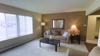 This single-floor space centers around a thoughtfully designed living room that combines comfort and functionality. The room features warm taupe-toned accent walls contrasted by lighter neutral paint, creating a welcoming and cohesive color palette throughout. Soft, plush wall-to-wall carpeting adds warmth underfoot and contributes to the overall cozy ambiance.

A large window fitted with vertical blinds allows ample natural light to fill the room, brightening the space during the day. A baseboard heating unit runs along the base of the window wall, providing a practical climate-control solution. The living area is well-proportioned, comfortably accommodating a cushioned sofa adorned with decorative pillows, a dark-finished rectangular coffee table, and additional seating.

Decorative elements include a framed wall mirror above the sofa, which enhances the sense of depth in the room, while two floor and table lamps provide warm, layered ambient lighting for evening use. The overall atmosphere is calm and inviting, with a neutral, versatile design that lends itself well to a variety of furnishing styles.