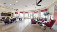 This single-floor community clubhouse is thoughtfully designed to offer a welcoming and versatile gathering space for residents. The main lounge area is generously proportioned, featuring a mix of slate tile and carpet flooring, tray ceilings, multiple ceiling fans, and recessed lighting that creates a bright and comfortable atmosphere. Large multi-pane windows dressed with rich burgundy valances line the walls, flooding the interior with natural light and offering views of the outdoor pool area. Seating throughout the lounge includes deep red leather wingback chairs with nailhead trim arranged around round tables, providing inviting conversation areas. A wall-mounted flat-screen television and framed artwork contribute to the refined yet relaxed ambiance. Elegant arched doorways connect the various areas of the clubhouse, including a dedicated billiards room equipped with a full-sized pool table, high counter seating, and additional windows overlooking the pool. The kitchen area is well-appointed with rich dark wood cabinetry, granite countertops, stainless steel appliances including a full-size refrigerator and microwave, and a coffee station, making it fully functional for events and everyday use. The overall design balances comfort and style, making this clubhouse an ideal hub for community activities.