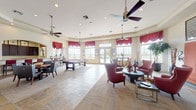 This single-floor community clubhouse is thoughtfully designed to offer a welcoming and versatile gathering space for residents. The main lounge area is generously proportioned, featuring a mix of slate tile and carpet flooring, tray ceilings, multiple ceiling fans, and recessed lighting that creates a bright and comfortable atmosphere. Large multi-pane windows dressed with rich burgundy valances line the walls, flooding the interior with natural light and offering views of the outdoor pool area. Seating throughout the lounge includes deep red leather wingback chairs with nailhead trim arranged around round tables, providing inviting conversation areas. A wall-mounted flat-screen television and framed artwork contribute to the refined yet relaxed ambiance. Elegant arched doorways connect the various areas of the clubhouse, including a dedicated billiards room equipped with a full-sized pool table, high counter seating, and additional windows overlooking the pool. The kitchen area is well-appointed with rich dark wood cabinetry, granite countertops, stainless steel appliances including a full-size refrigerator and microwave, and a coffee station, making it fully functional for events and everyday use. The overall design balances comfort and style, making this clubhouse an ideal hub for community activities.