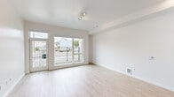 This single-floor unit offers a clean, contemporary design with a bright and minimalist aesthetic throughout. The open-concept living area is bathed in natural light, courtesy of large sliding glass doors and expansive windows that provide views of the surrounding neighborhood and open onto a railed balcony. Light wood-tone flooring runs throughout the space, complementing the crisp white walls and creating a warm yet modern atmosphere. Track lighting overhead adds a functional and stylish touch to the living area.

The kitchen is efficiently designed along a single wall and features wood-grain cabinetry that adds texture and warmth to the otherwise neutral palette. Stainless steel appliances — including a refrigerator, electric range, dishwasher, and microwave — are neatly integrated alongside a white tile backsplash and a simple undermount sink. Recessed lighting above the kitchen enhances the clean, polished look. A sliding closet is conveniently located just off the kitchen corridor, providing practical storage.

The bathroom is finished in all-white surfaces with neutral tile flooring, offering a clean and functional space complete with accessibility-friendly grab bars. Overall, the unit presents a streamlined, move-in-ready environment with thoughtful design details and ample natural light throughout.